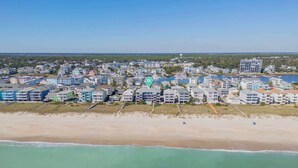 Ovenudblik over et kystnært boligområde med ejendomme ved stranden, en lysthavn og en klar himmel.