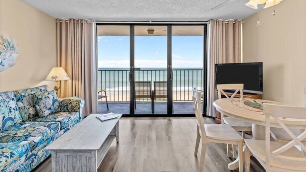 Una habitación con sofá, mesa de centro, televisor, mesa de comedor con sillas, ventilador de techo y vista a la playa a través de una puerta corredera.