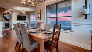 A dining area with a wooden table set for four, surrounded by wicker chairs. A large window with a view of a sunset and mountains is visible. - Lake View @ Hollows Resort: Pools, Pets, Balconies (Jonestown)