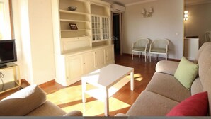 Een woonkamer met een bank, een salontafel en een tv-meubel. Er zijn twee stoelen en een kast met planken.