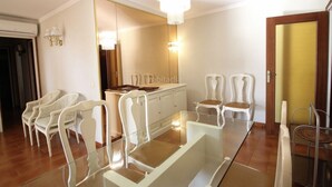 Een moderne woonkamer met een glazen eettafel, witte stoelen, een grote spiegel en een kroonluchter.
