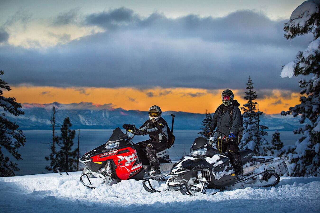 Two snowmobiles with riders in a snowy landscape with mountains in the background.