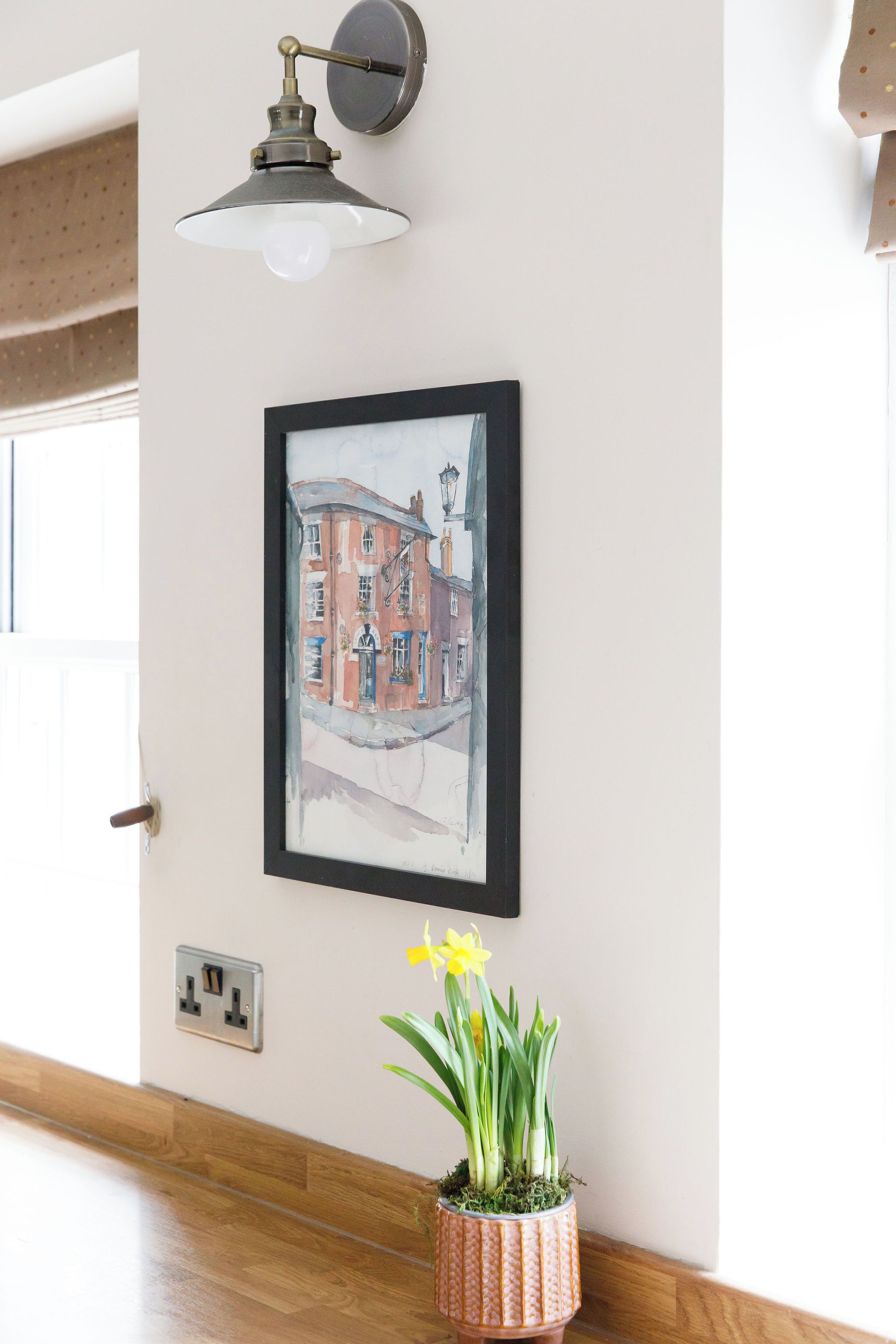 A framed watercolor painting of a building, a potted plant with yellow flowers, and a wall-mounted light fixture.