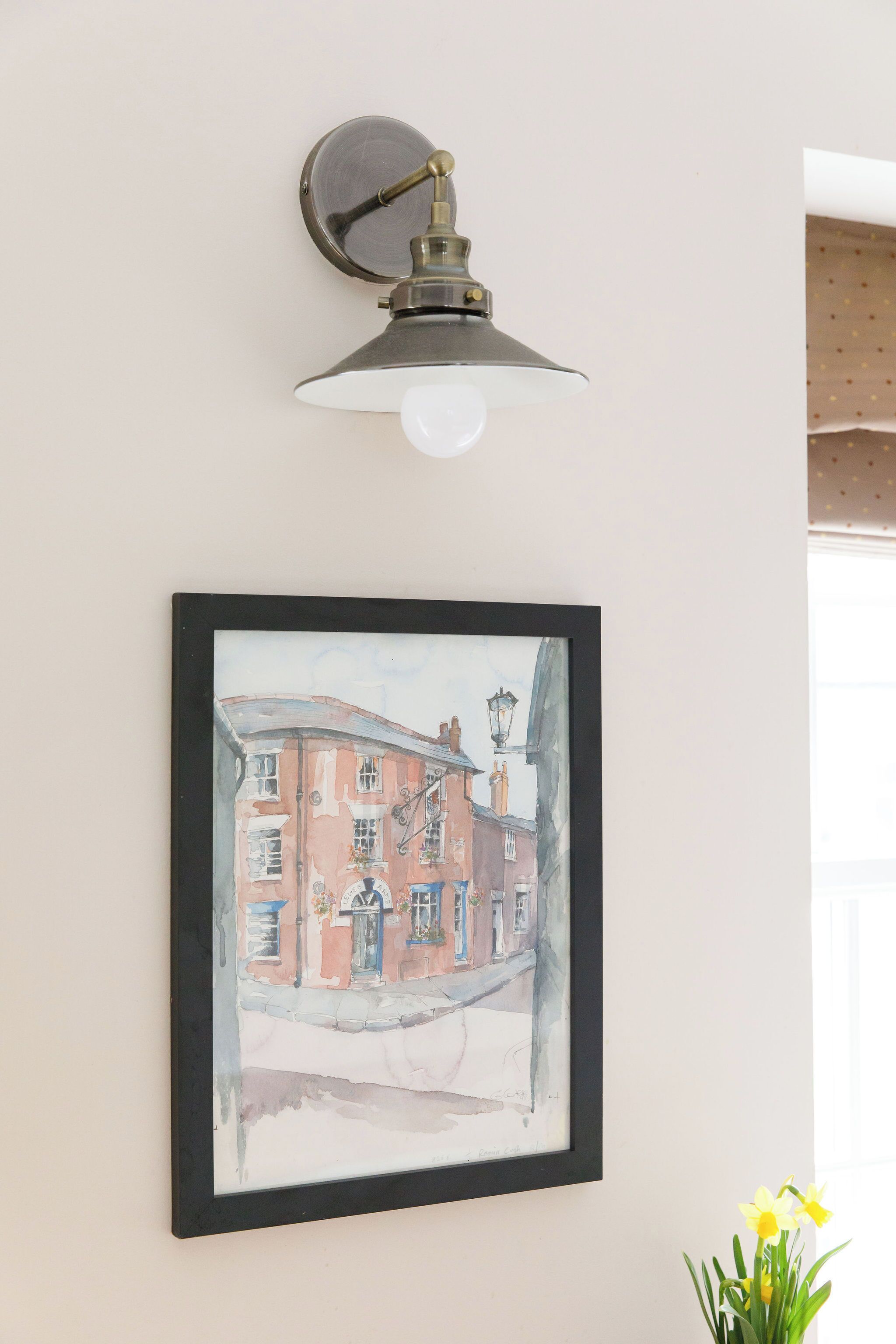 Ein gerahmtes Aquarellgemälde eines Gebäudes an einer Wand, eine Wandleuchte und eine Vase mit Blumen.