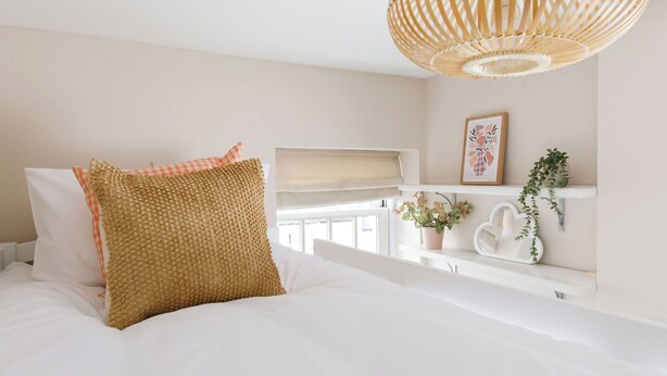 Een slaapkamer met een bed, een raam met zonwering, een nachtkastje met een plant, een wandplank met decoratieve objecten en een hangende lamp.