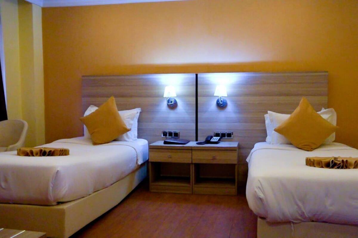 A hotel room with two single beds, each with a pillow and a bedside table with a lamp and a phone.