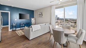 A modern living room with a sofa, dining table, and chairs, a flat-screen TV, and a city view through large windows. - The Malibu Suite Penthouse The Gulch
 (Nashville)