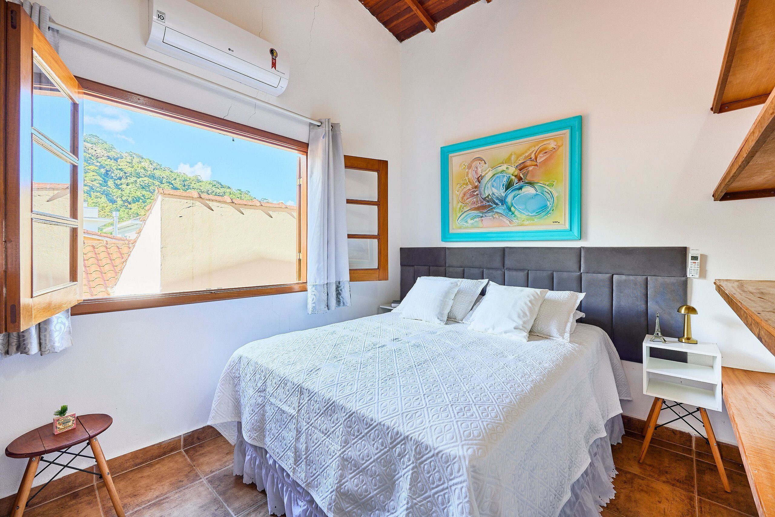 A bedroom with a bed, a window, a painting, and a bedside table.