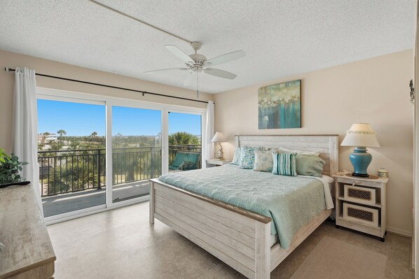 A bedroom with a bed, a nightstand, a ceiling fan, a window with a view, and a painting on the wall. - Parrot Party - Ocean House 319 (St. Augustine)