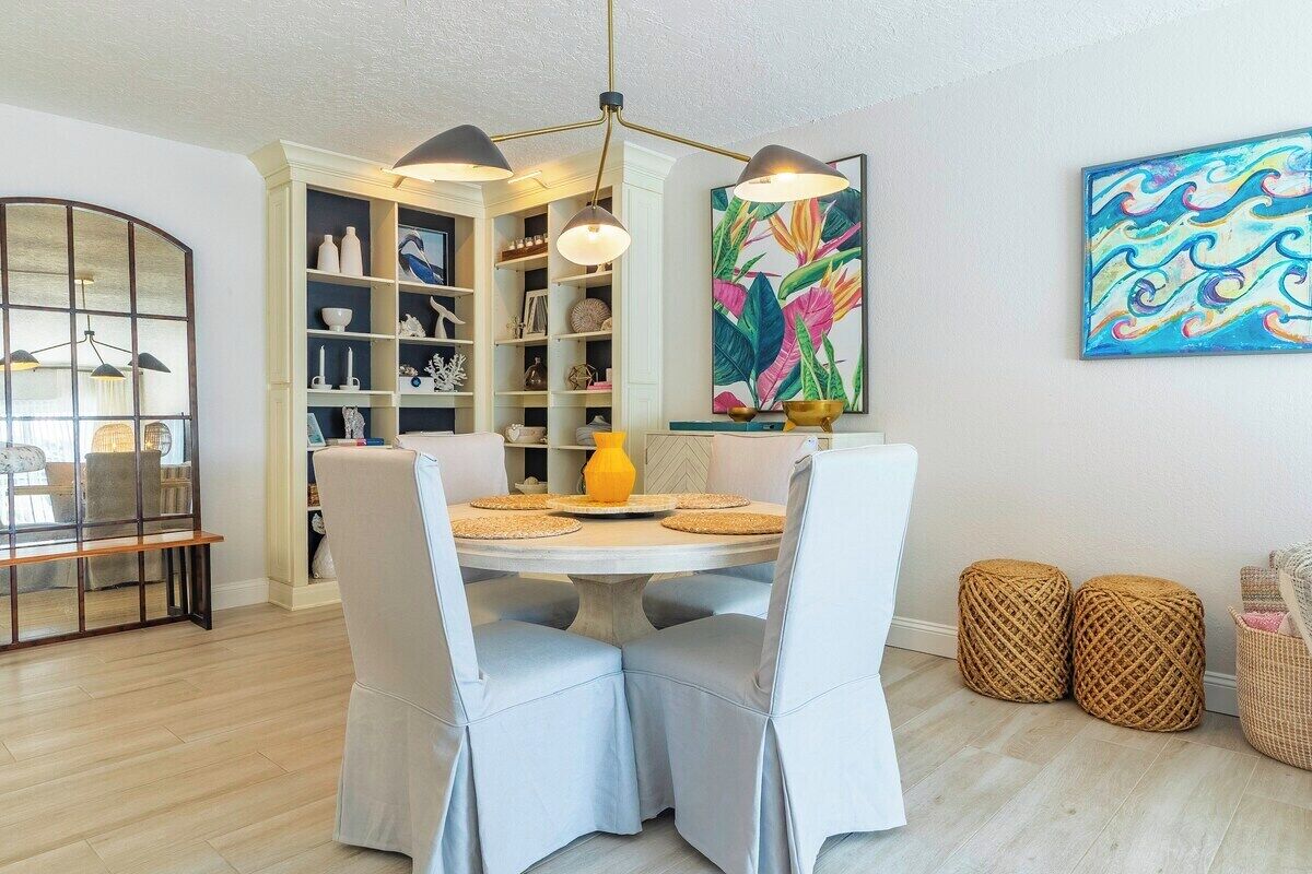A dining area with a round table, two chairs, a yellow vase, and a bookshelf with decorative items.