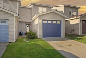 Une maison à deux étages avec une porte de garage bleue, un petit jardin devant la maison avec une pelouse et des dalles, et une antenne parabolique sur le toit.