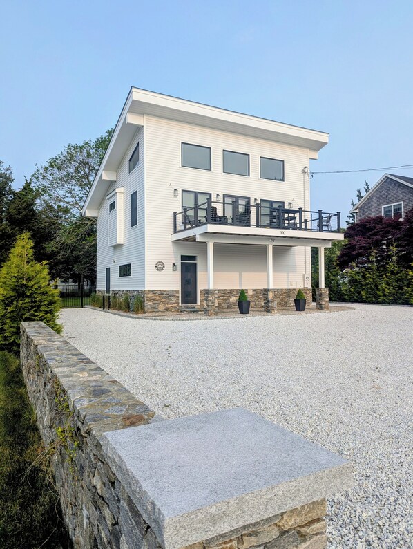 A modern two-story house with a covered front porch, a stone retaining wall, and a gravel driveway. - Modern Beach House Serenity (Westerly)