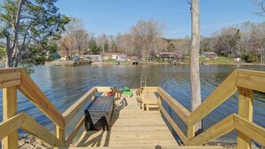 A wooden dock extending over a calm lake with a table and chairs, surrounded by trees and houses in the background.