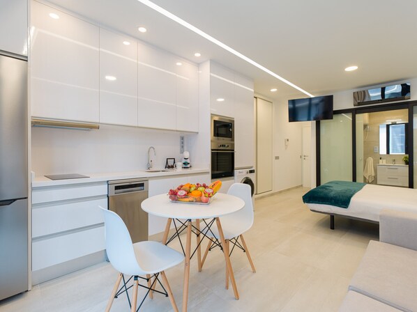 A modern kitchen with white cabinets, a dining area with a white table and chairs, and a living area with a sofa and a TV.