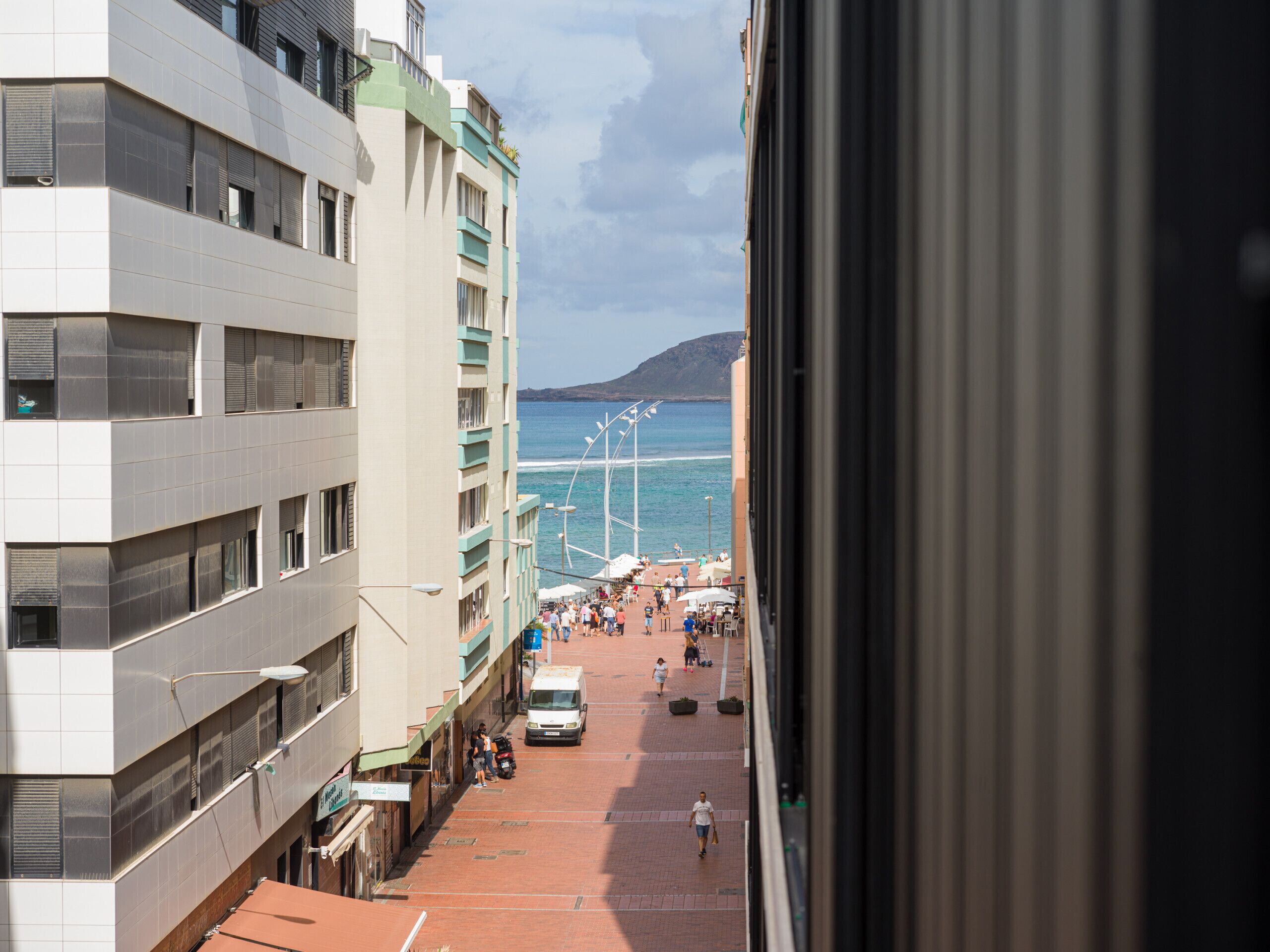 Vista de una calle costera con edificios, una furgoneta estacionada y gente caminando.