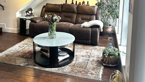 A cozy living room with a brown leather sofa, a round coffee table with a vase, a patterned rug, and a staircase with a sloped ceiling. - Stunning and Luxurious Riverfront home for 6 with two king beds. Pet Friendly! (Westwood)