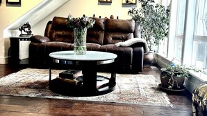 A living room with a brown leather sofa, a round coffee table with a vase, wooden flooring, and a staircase with wall-mounted shelves. - Stunning and Luxurious Riverfront home for 6 with two king beds. Pet Friendly! (Westwood)