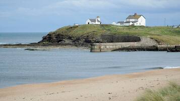A coastal scene with a sandy beach, a rocky outcrop, and a white building with a tower on a grassy hill.