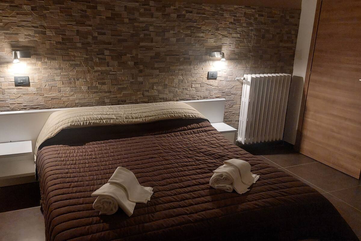 Une chambre moderne avec une tête de lit à motifs de pierre, un lit bien fait avec une couverture marron, et deux lampes fixées au mur.