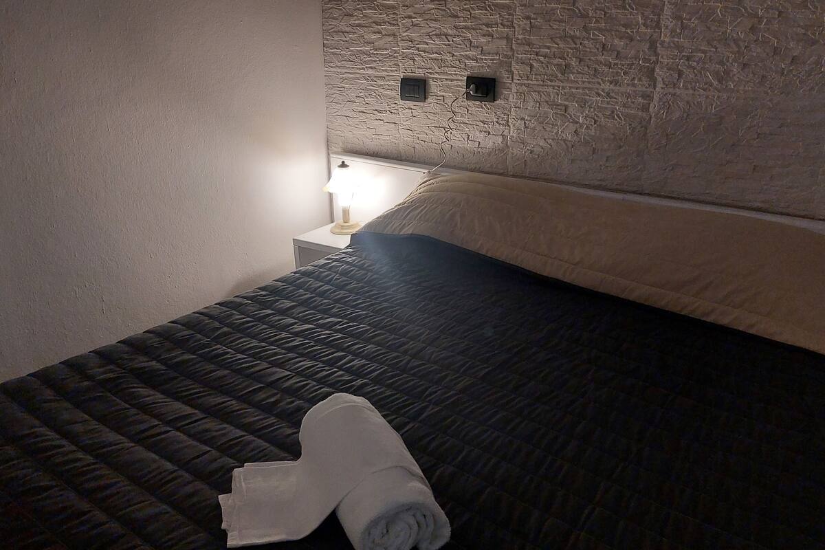 Un lit recouvert d’une courtepointe matelassée, une serviette enroulée et une table de chevet avec une lampe.