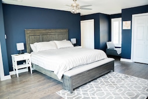 A bedroom with a large bed, a nightstand, a bench, a ceiling fan, and a window with blinds.