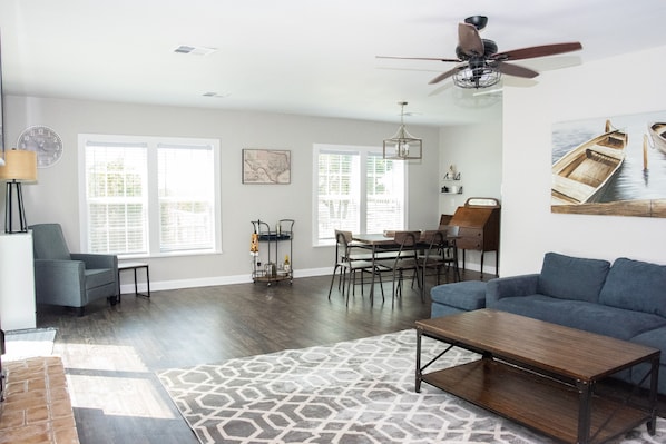 A spacious living room with a large sofa, a coffee table, and a dining area with a table and chairs. There is a ceiling fan, a chandelier, and a large window with blinds.