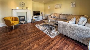 Een woonkamer met een bank, fauteuil, televisie, open haard en houten vloer.