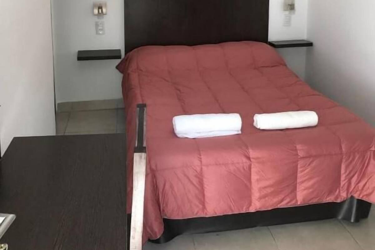 A hotel room with a bed, a bedside table, a wall-mounted light, and a small artwork above the bed.