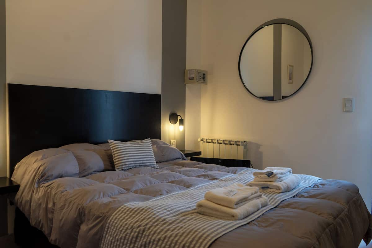 A neatly made bed with a striped blanket and folded towels, a round mirror on the wall, and a nightstand with a lamp.