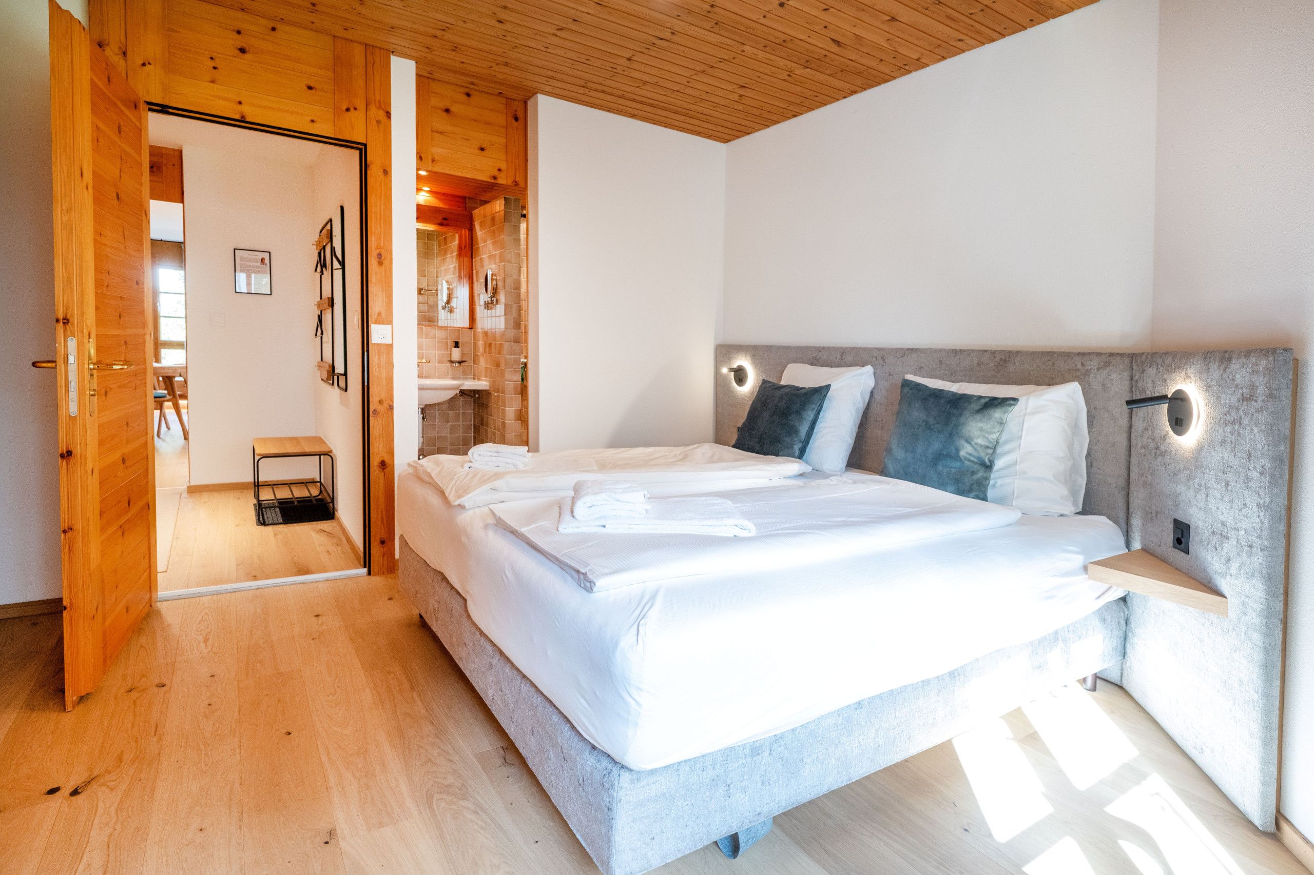Un dormitorio con techo de madera, una cama grande con ropa blanca, una mesita de noche y un banco empotrado.