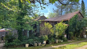 A rustic wooden house with a tiled roof, surrounded by lush greenery and a gravel path.