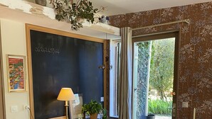 A room with a wooden desk, a red chair, a blackboard, a window with curtains, and a view of greenery outside. - B&B Les Truffières in the heart of nature (Trémolat)