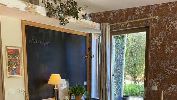 A room with a wooden desk, a red chair, a blackboard, a window with curtains, and a view of greenery outside.