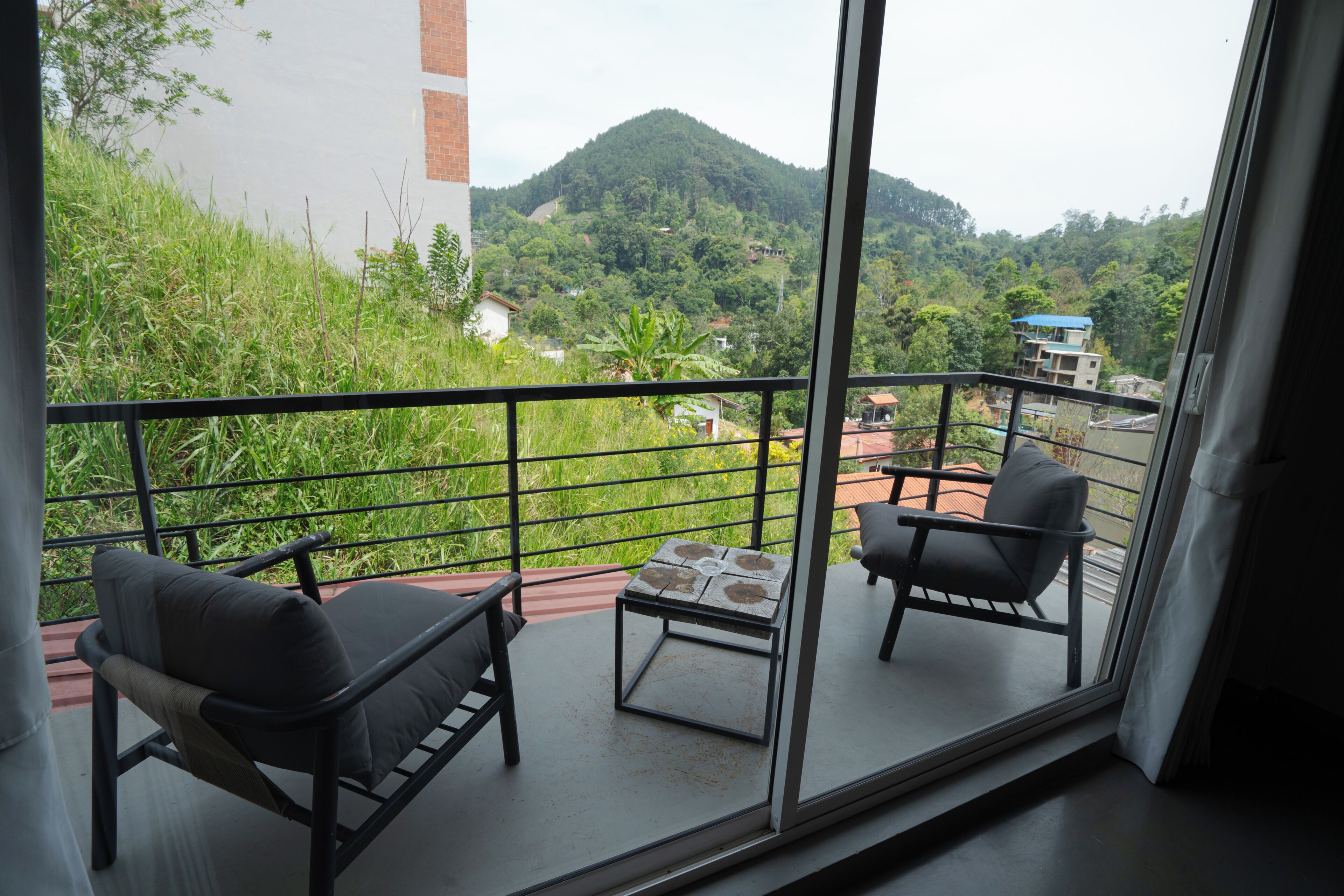 A balcony with two chairs and a small table, overlooking a lush green hillside with buildings in the distance.