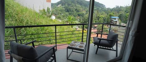 A balcony with two chairs and a small table, overlooking a lush green hillside with buildings in the distance.