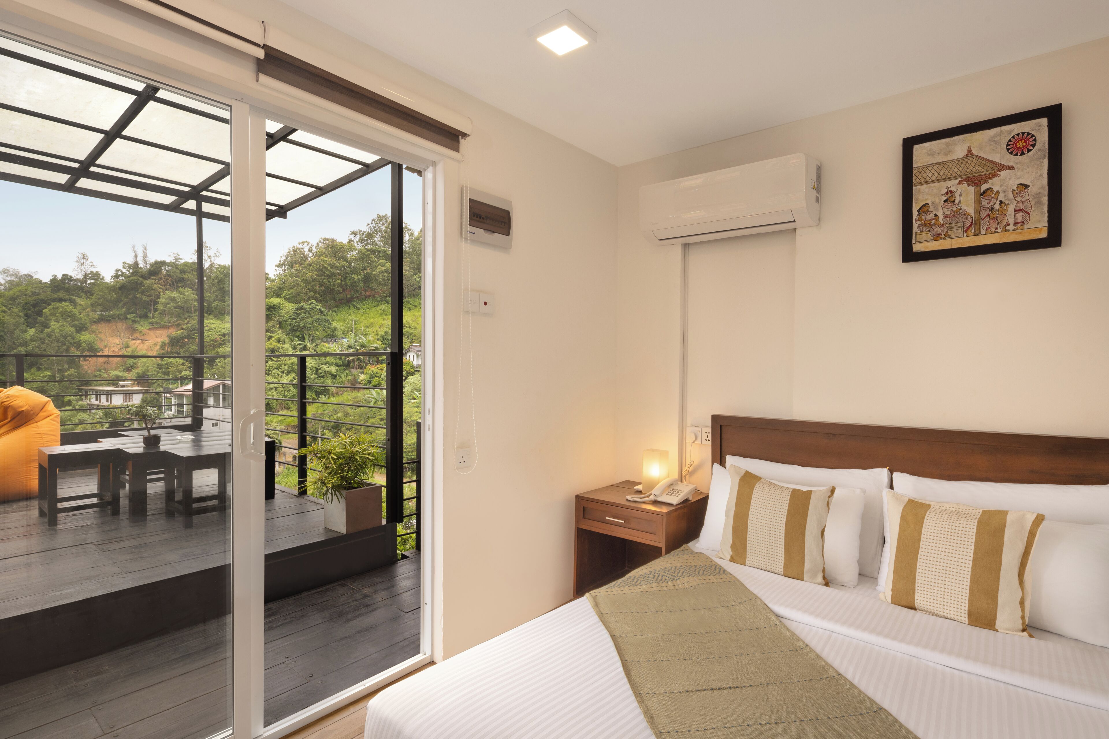 A hotel room with a bed, a bedside table, a nightstand, a lamp, a painting on the wall, and a balcony with a table and chairs.