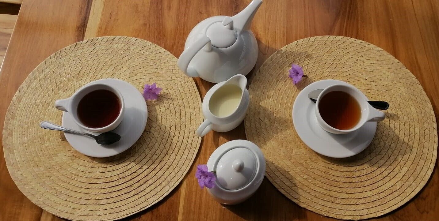 A table with two cups of tea, a teapot, and a small jug on woven placemats.