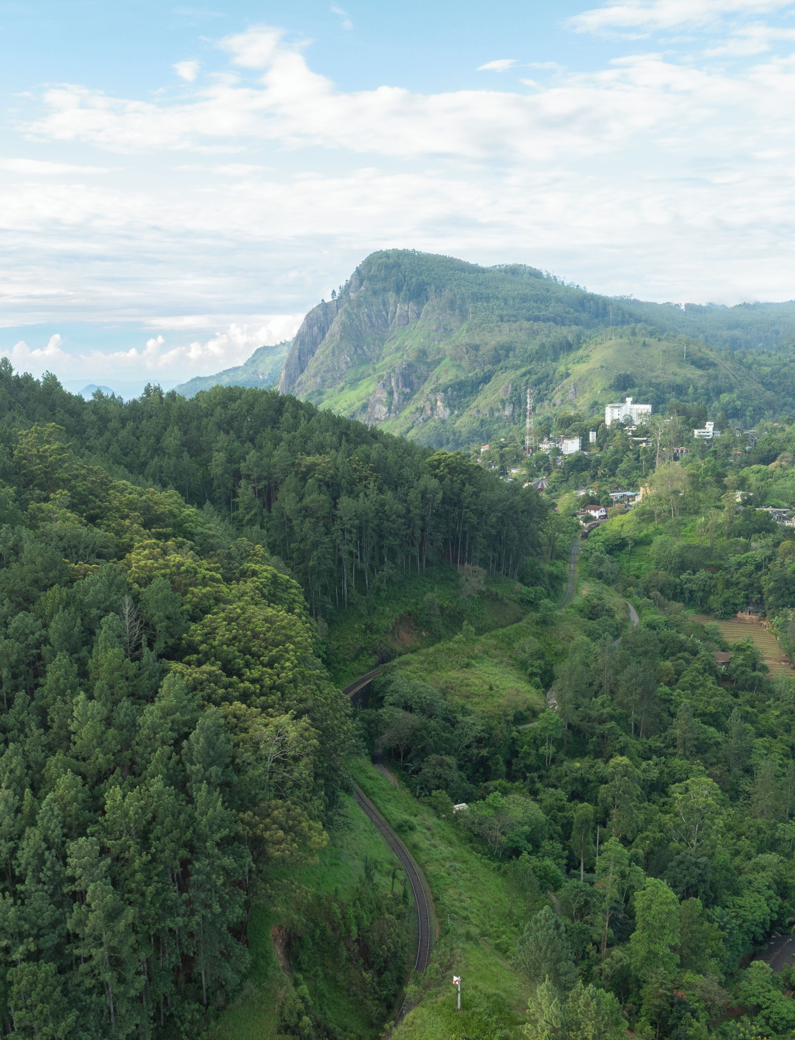 A lush green valley with a winding road, a prominent mountain, and scattered buildings.
