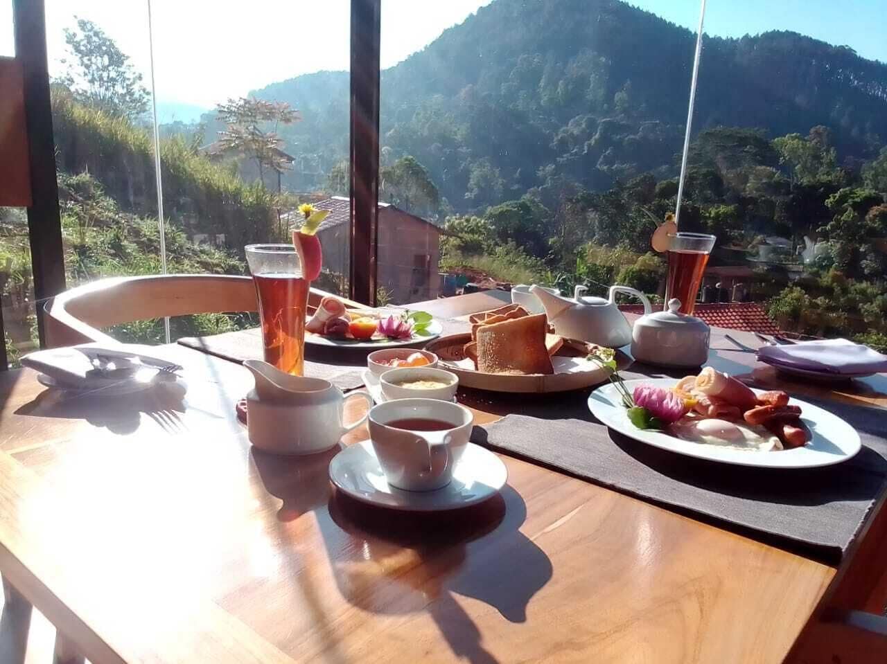A breakfast table with various dishes, cups, and a teapot, set against a scenic mountain backdrop.