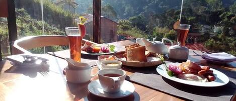 Mesa de café da manhã com diversos pratos, xícaras e uma chaleira, ao fundo uma paisagem montanhosa.