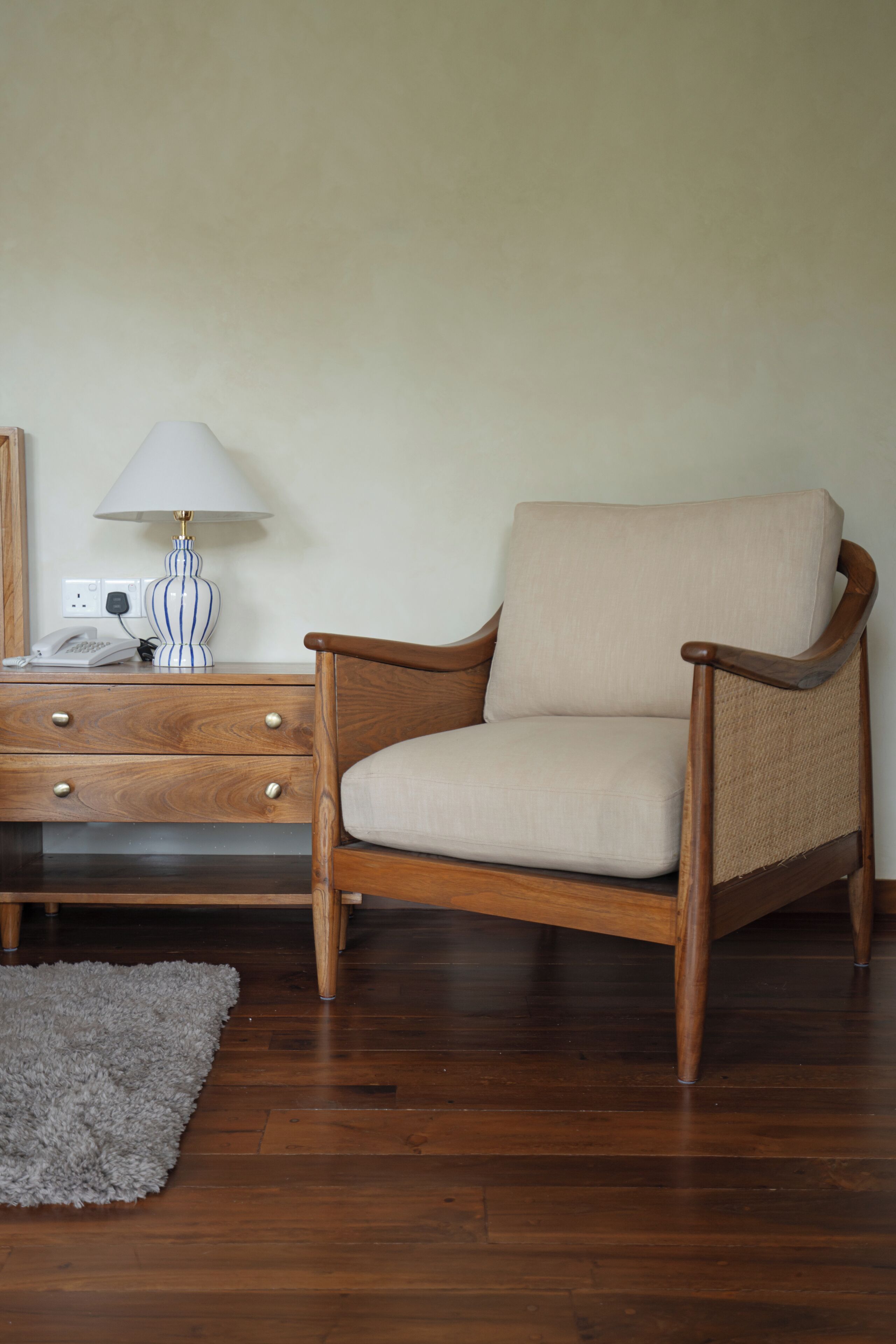 A wooden chair with a cushion, a lamp, a telephone, and a small rug on a wooden floor.