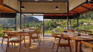 An open dining area with wooden tables and chairs, large windows offering a view of a lush green landscape. - The Cubes Ella  (Ella)