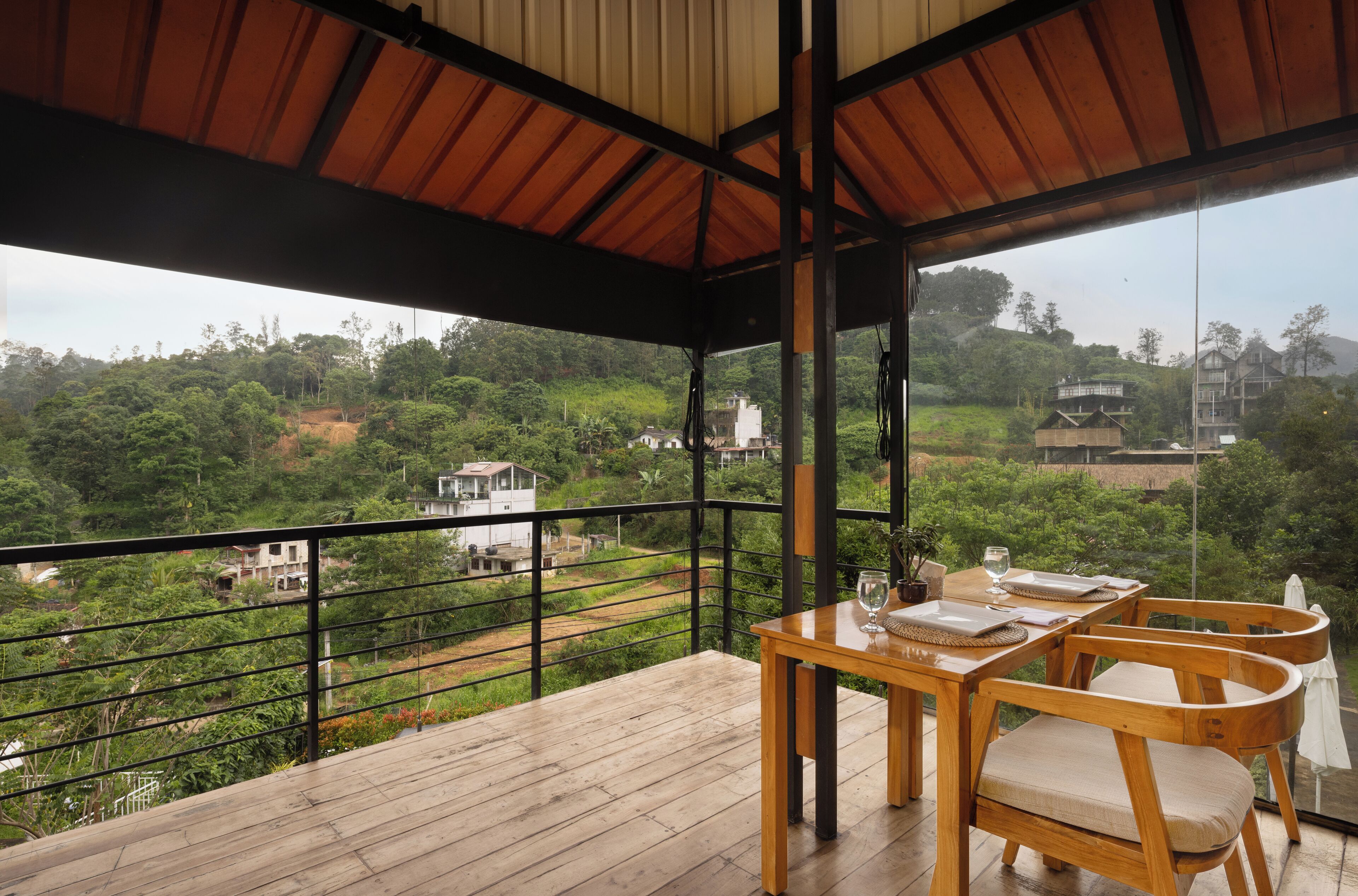 A wooden deck with a table and chairs, overlooking a lush green landscape with buildings in the distance.