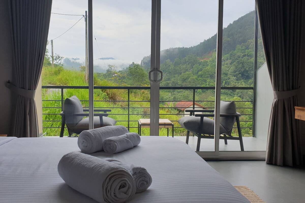A hotel room with a large bed, two chairs, a small table, and a balcony with a view of green hills.