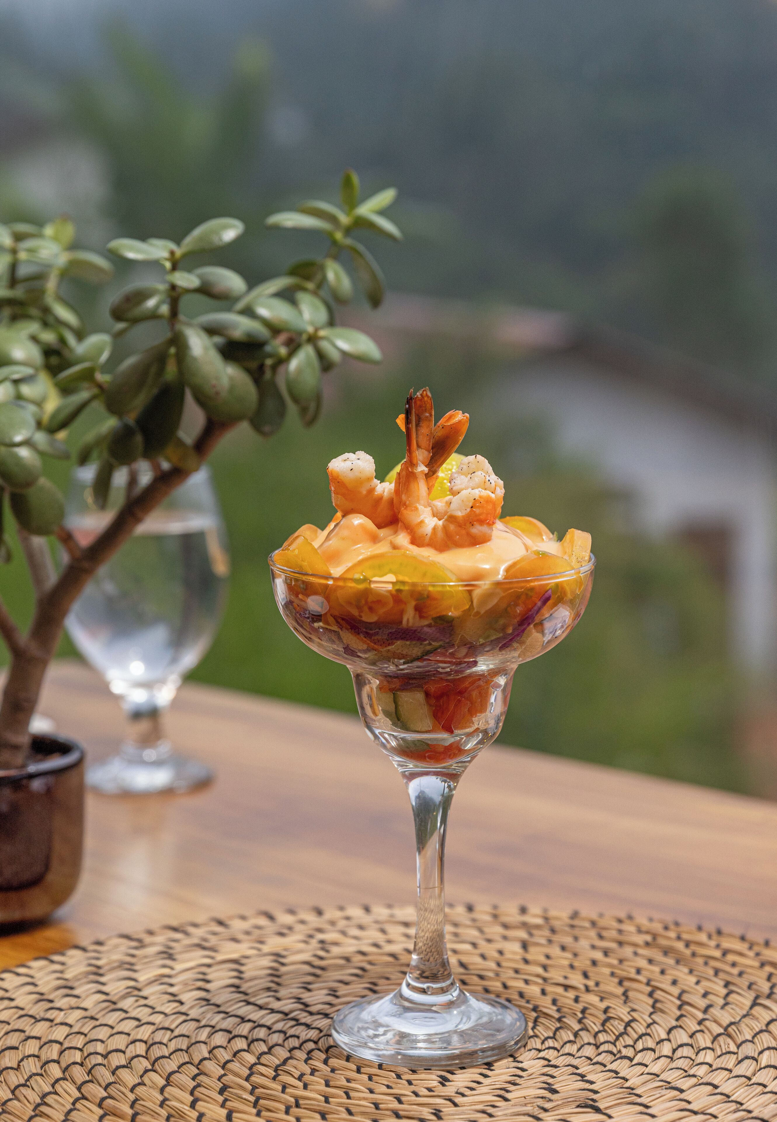 A dessert in a glass bowl on a woven placemat, with a potted plant and a wine glass in the background.