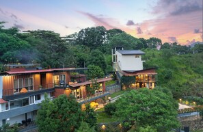 A modern multi-story house with a balcony, surrounded by lush greenery and a scenic sunset view. - The Cubes Ella (Ella)