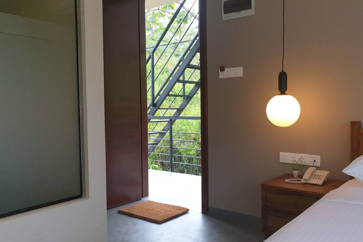 A bedroom with a bed, bedside table with a phone, a wall-mounted light, a door leading outside, and a small rug.