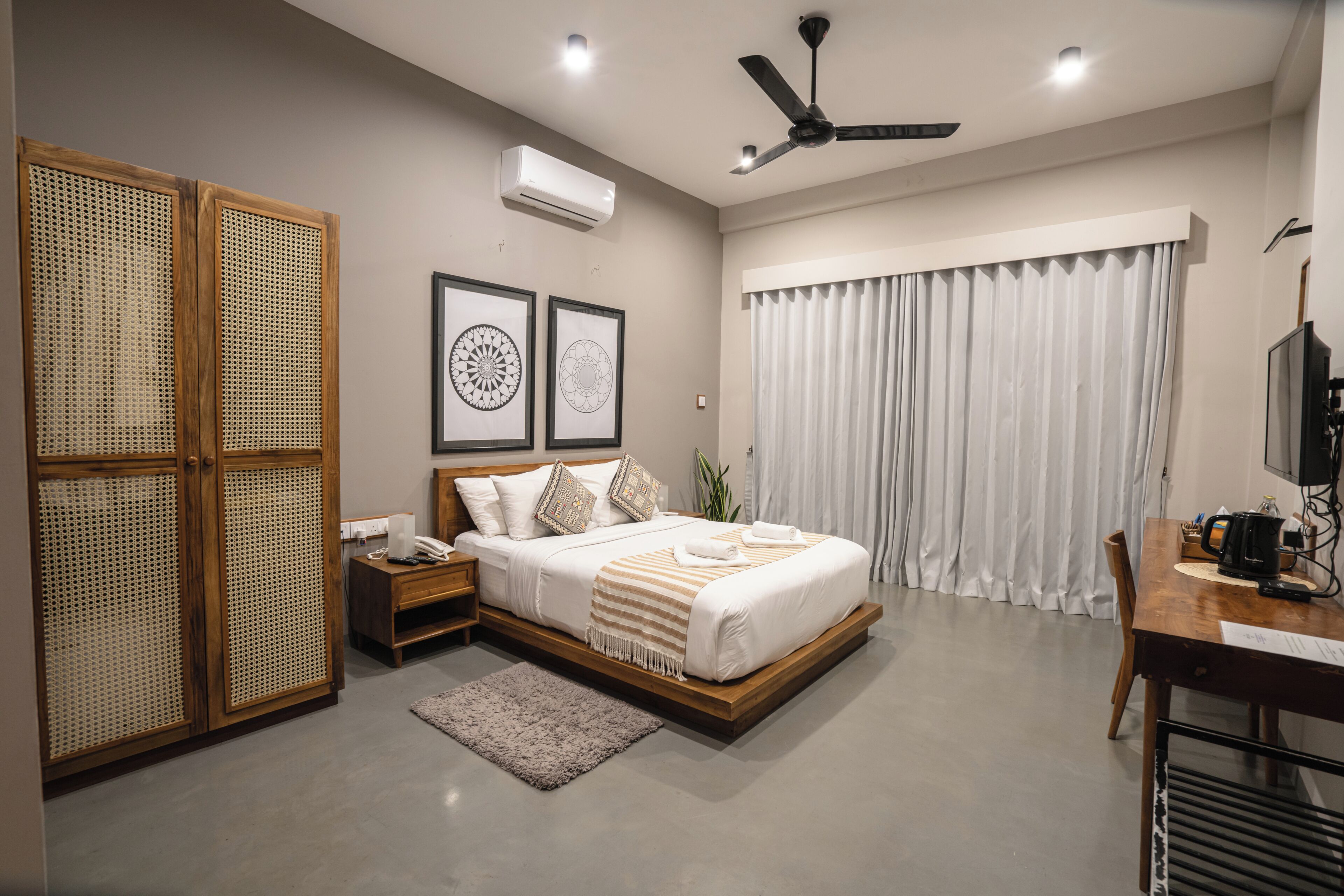 A bedroom with a bed, bedside tables, a desk, a ceiling fan, and a large window with curtains.