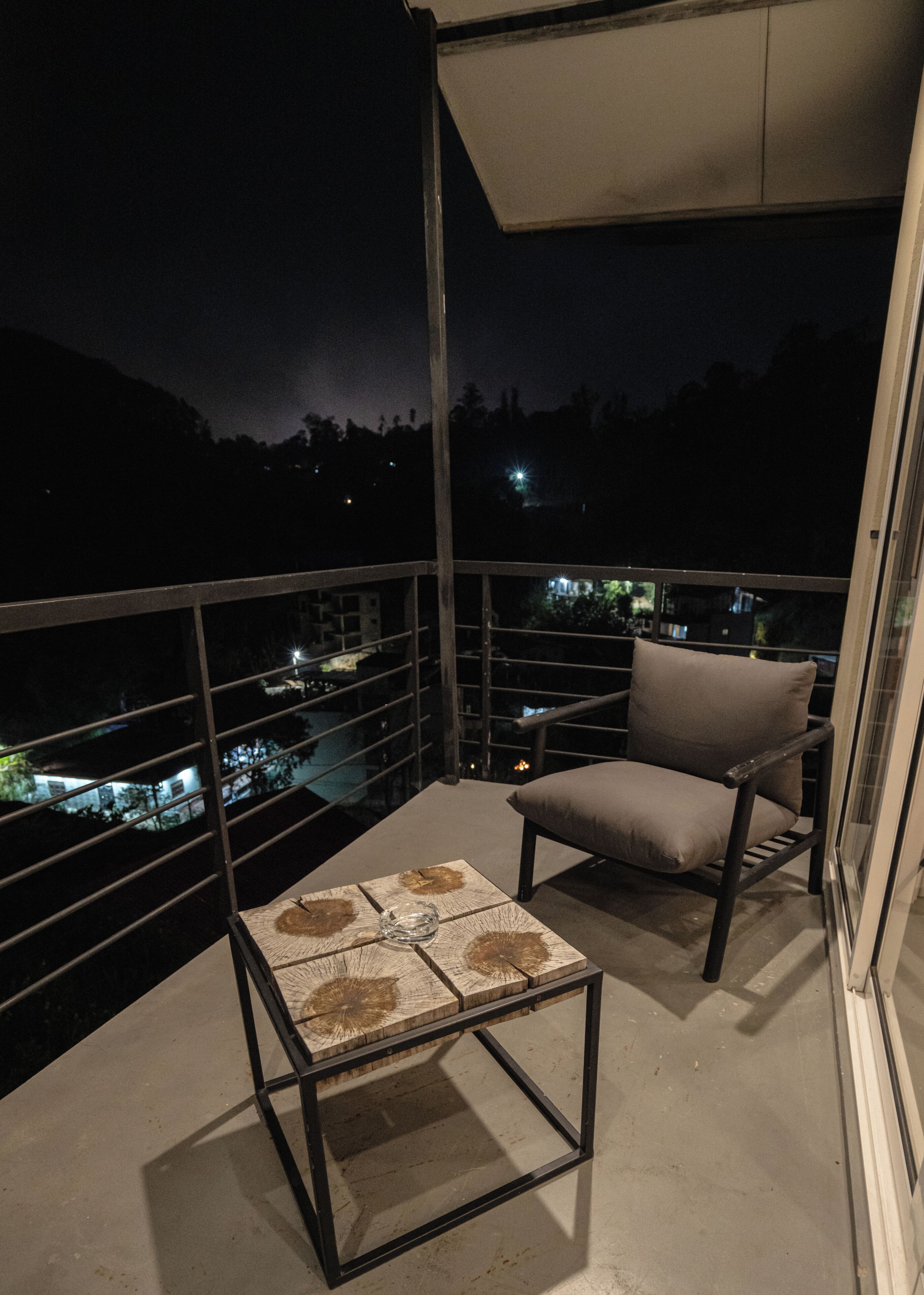 A balcony with a chair and a table, overlooking a cityscape at night.