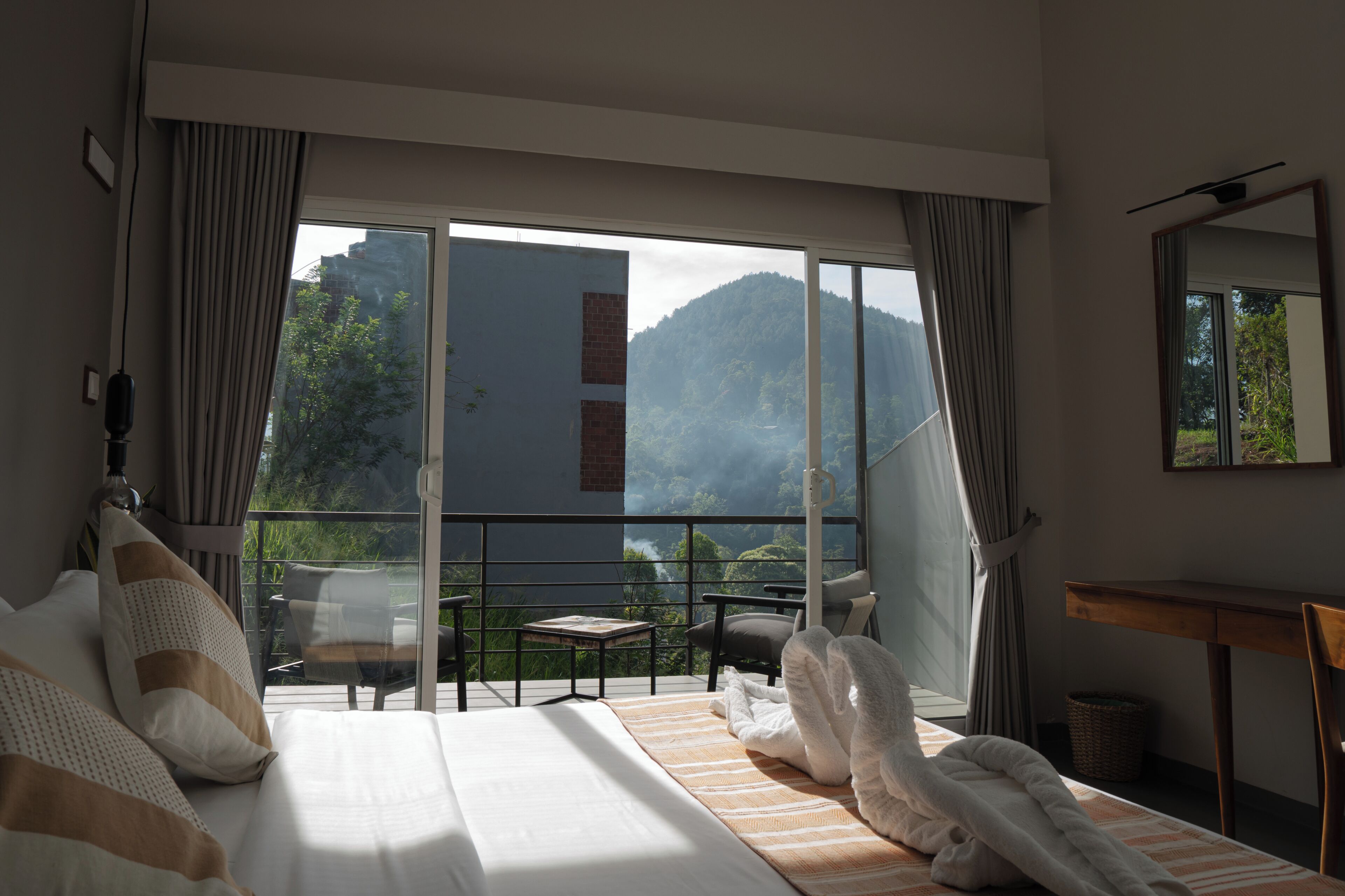 A hotel room with a bed, a small table, a chair, and a balcony with a view of a mountain.